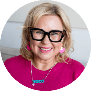 Close-up portrait of a smiling blonde woman wearing black-framed glasses, a pink sweater, pink earrings and a turquoise pendant necklace.