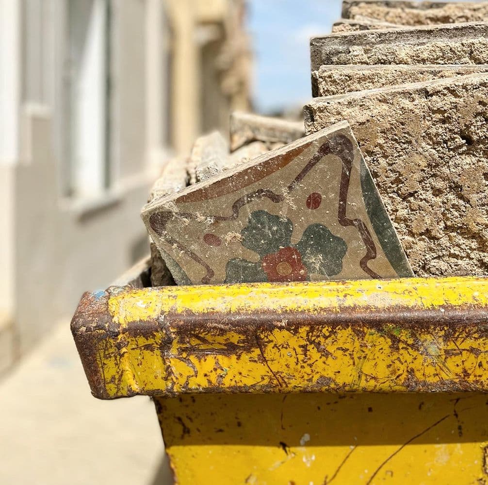 A colorful, patterned tile sits among weathered, broken tiles in a yellow industrial container on a sunny street.