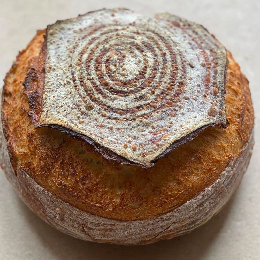Loaf of round sourdough bread with a crisp, golden crust and a spiral pattern on top, placed on a light surface.