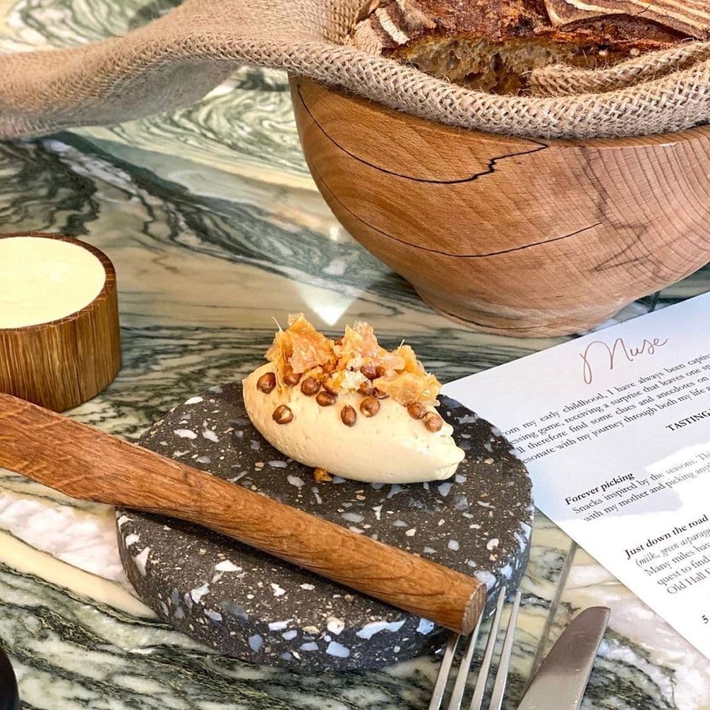 A piece of butter topped with grains on a stone plate, a wooden bread bowl, menu, and a wooden butter container on a marble table.