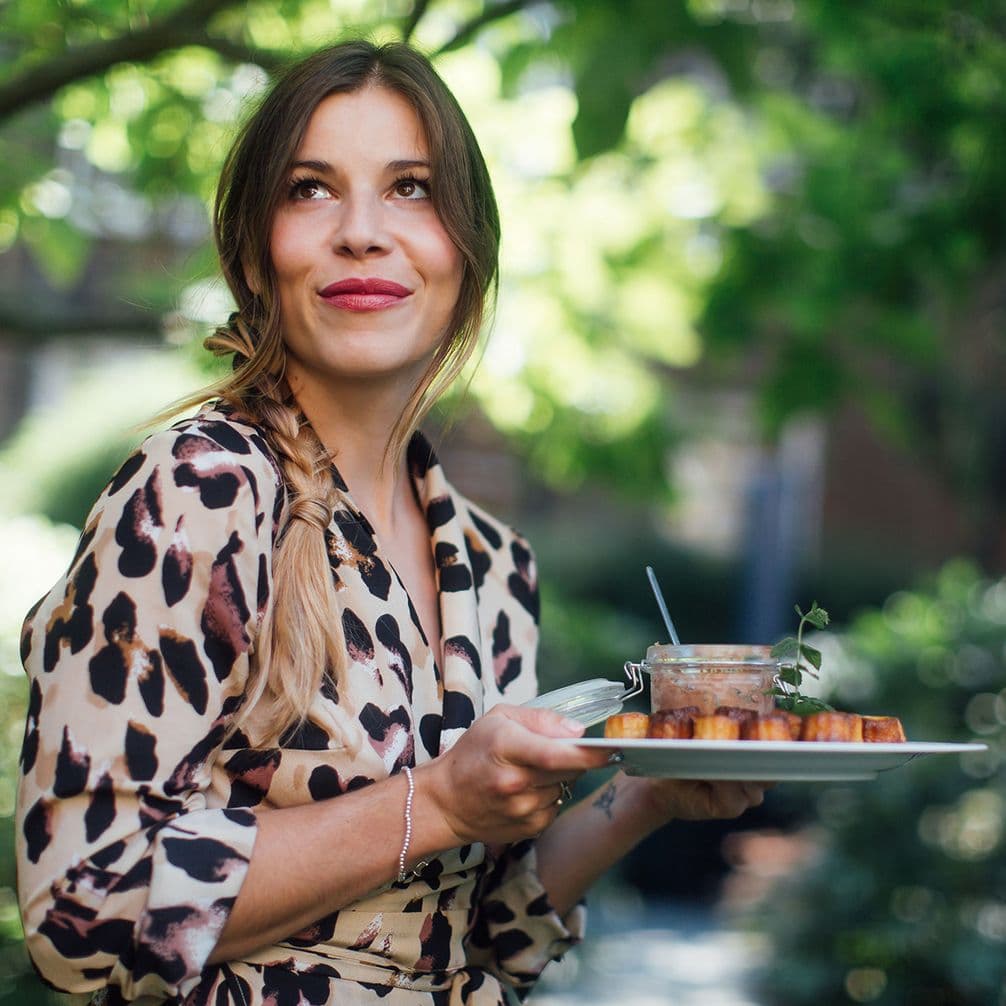A woman in a leopard-print dress holds a plate with food, smiling confidently outdoors, surrounded by greenery.
