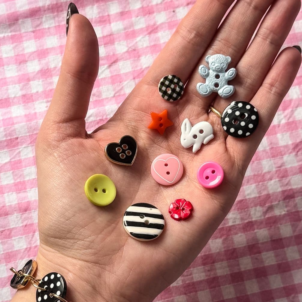 Open hand holding assorted colorful novelty buttons (heart, bunny, teddy, star, striped, polka-dot) on pink gingham background.