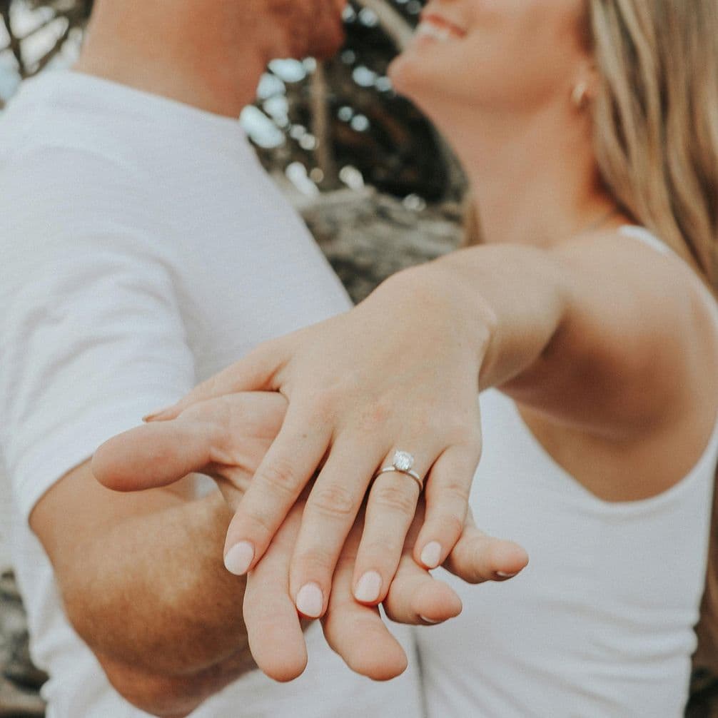 Couple smiling and embracing, woman extending hand to show engagement ring. Both wearing white tops, with blurred natural background.