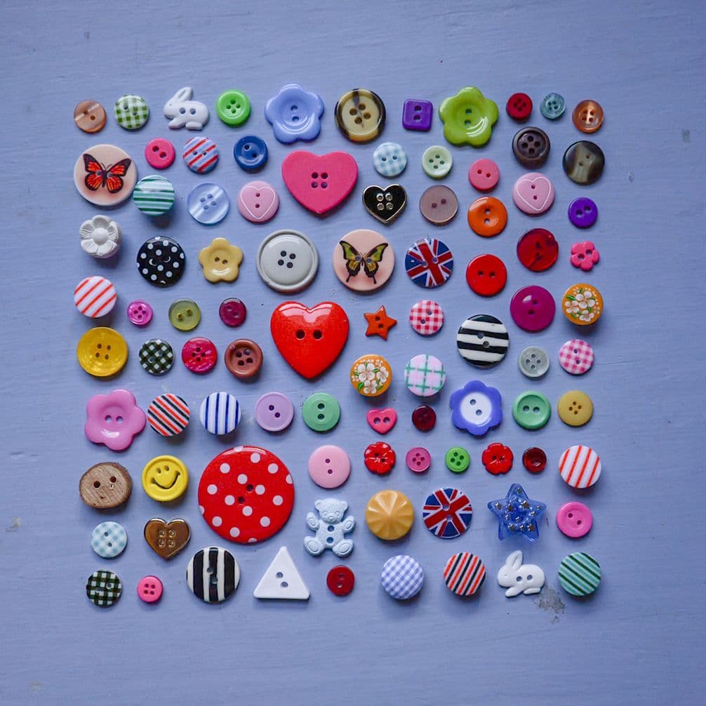 Assorted colorful buttons and novelty charms (hearts, flowers, flags, stars, animals) arranged in a neat square on a lavender background.