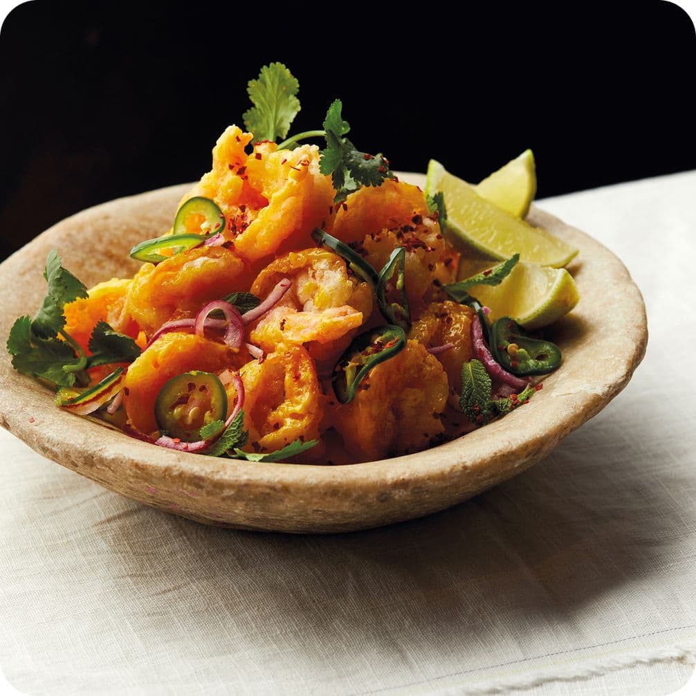 Bowl of crispy battered shrimp fritters garnished with jalapeño slices, pickled red onion, cilantro leaves and lime wedges.