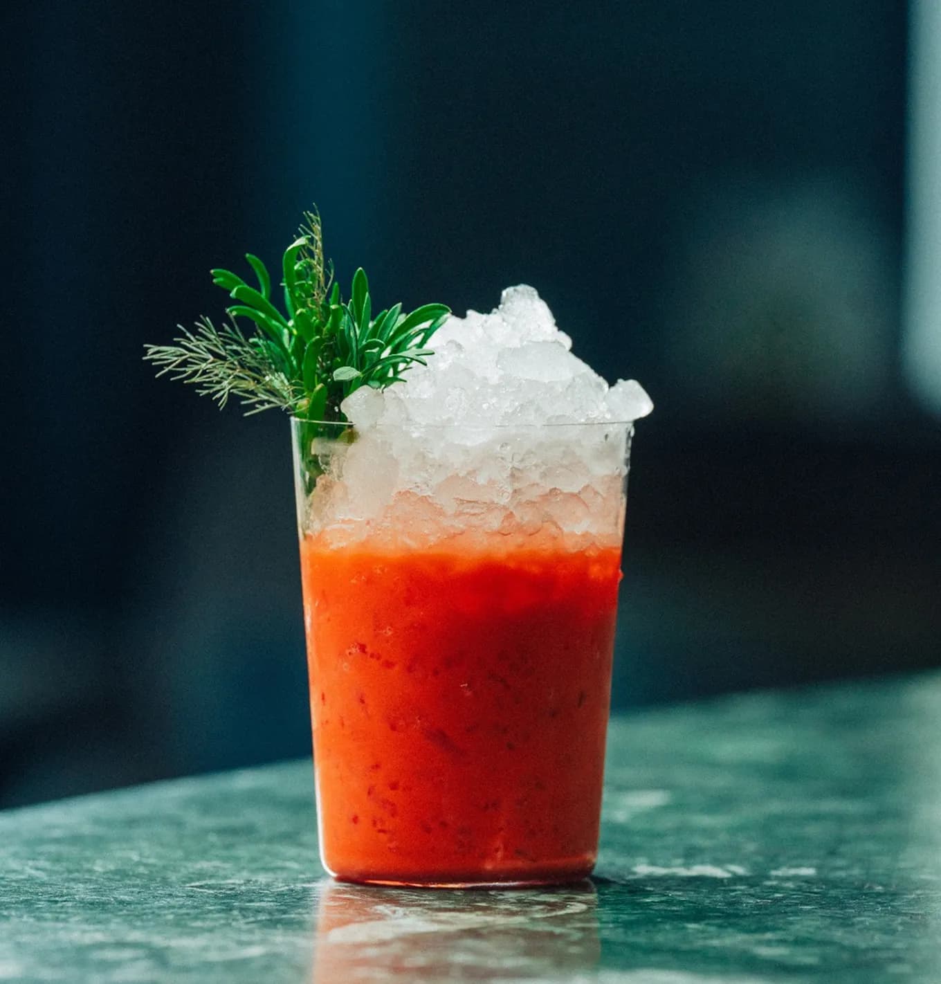 A vibrant red drink garnished with crushed ice and fresh greens, served in a clear cup on a marbled surface.