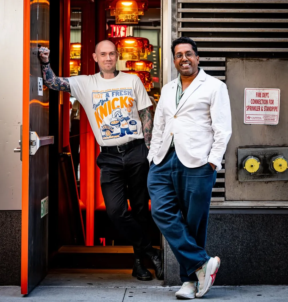 Two men standing outside a building; one is wearing a Knicks t-shirt, the other is in a white blazer. Both are smiling, with an industrial background.