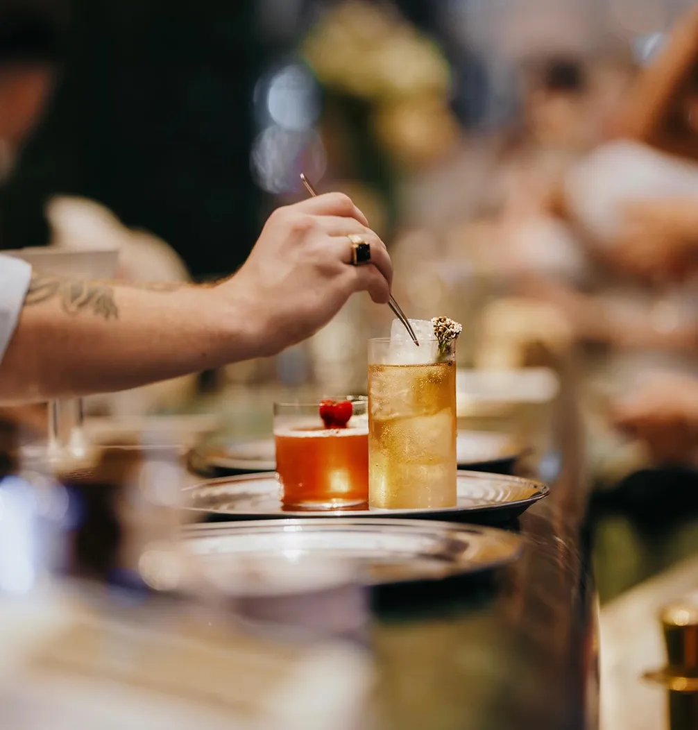 A hand stirring a golden cocktail with ice in a tall glass at a bar, next to a red drink with a berry garnish on a metallic tray.