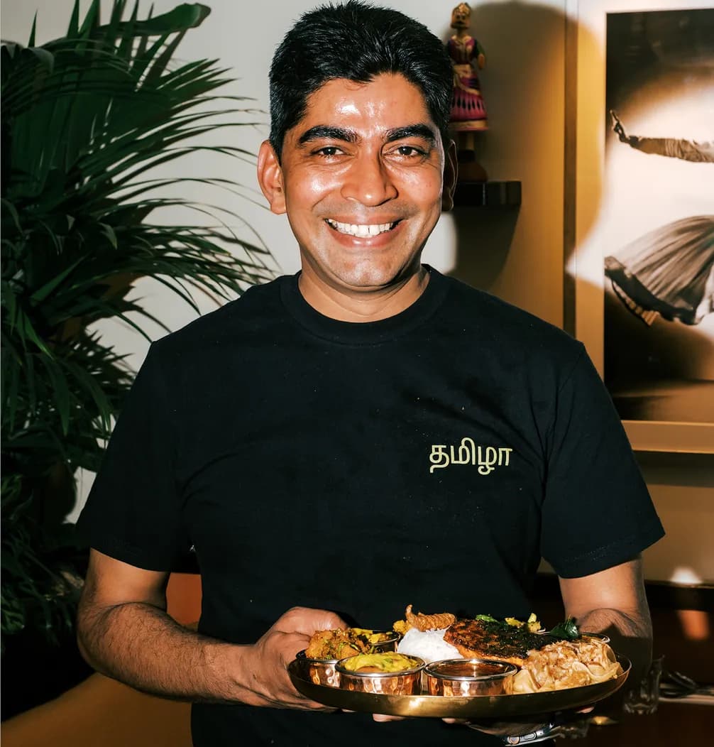 A smiling man holds a traditional Indian thali, wearing a black shirt with Tamil script, standing in a warmly lit room with plants and decor.