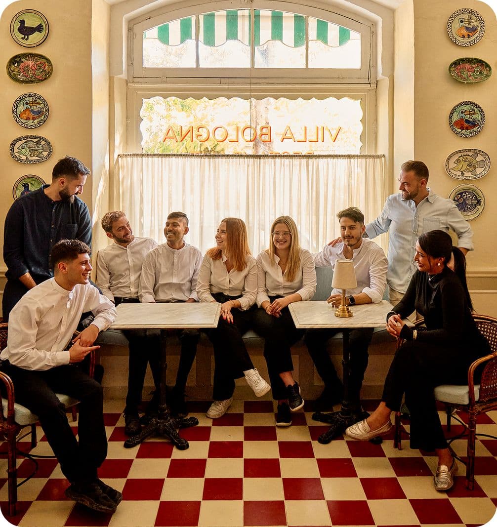 Nine people in white and dark attire chatting around cafe tables under a window sign "VILLA BOLOGNA", red-and-white checkered floor and decorative plates.