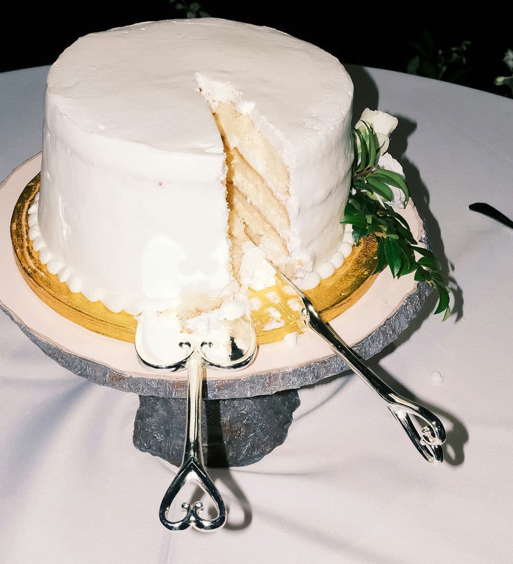 A white three-layer cake with a slice removed, adorned with white flowers and heart-shaped cake servers on a gold base, displayed on a table.