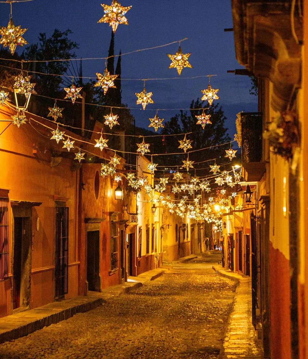 A cobblestone street at dusk, adorned with hanging star-shaped lights, lined with colorful buildings and warm street lamps.