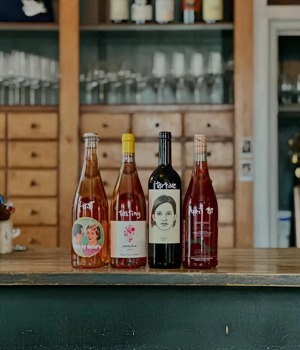 Four bottles of wine with artistic labels lined up on a wooden counter in a rustic room with shelves and glassware in the background.