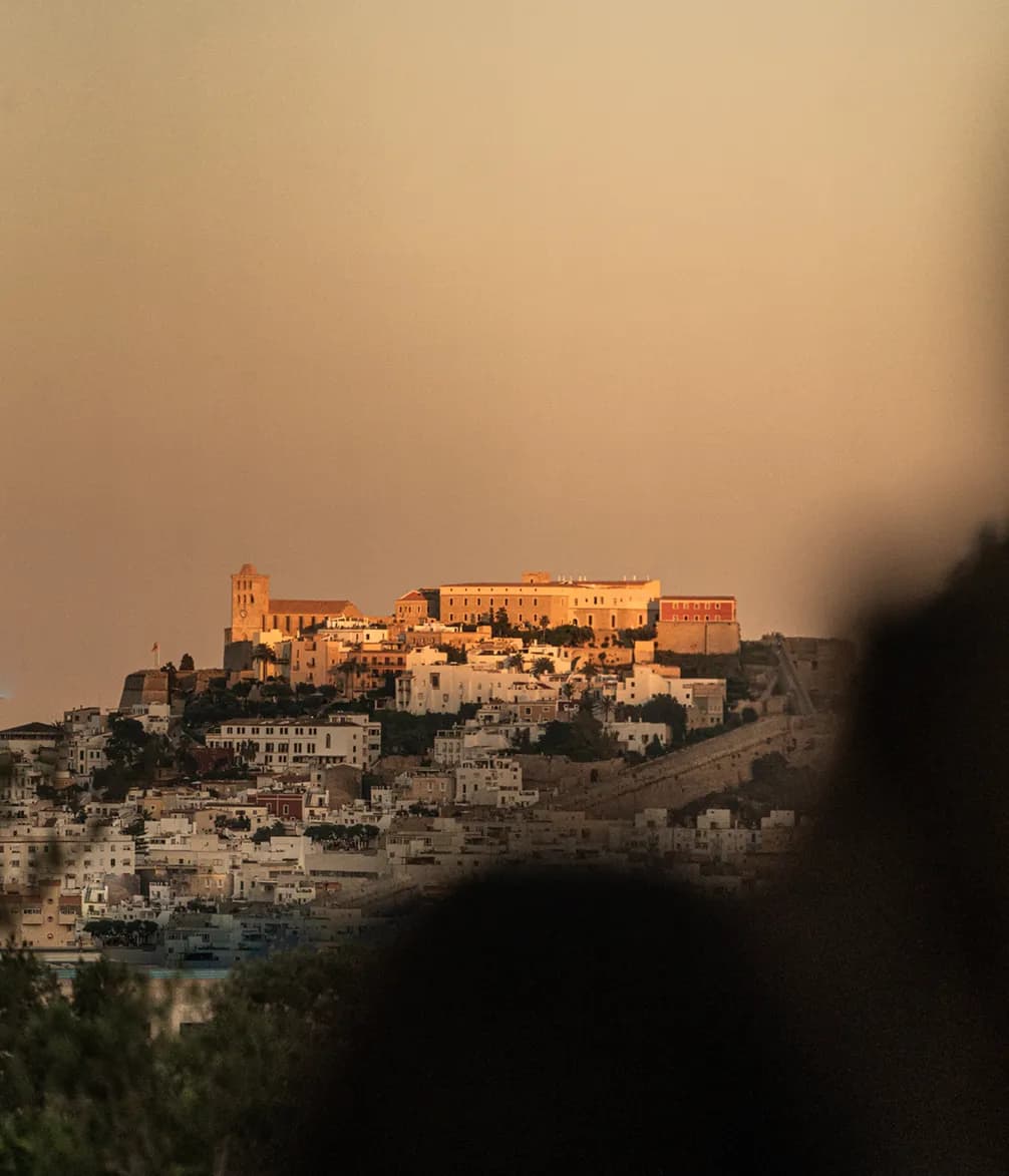 Hilltop cityscape at sunset with a historic building illuminated in golden light, surrounded by smaller white structures and a blurred foreground.