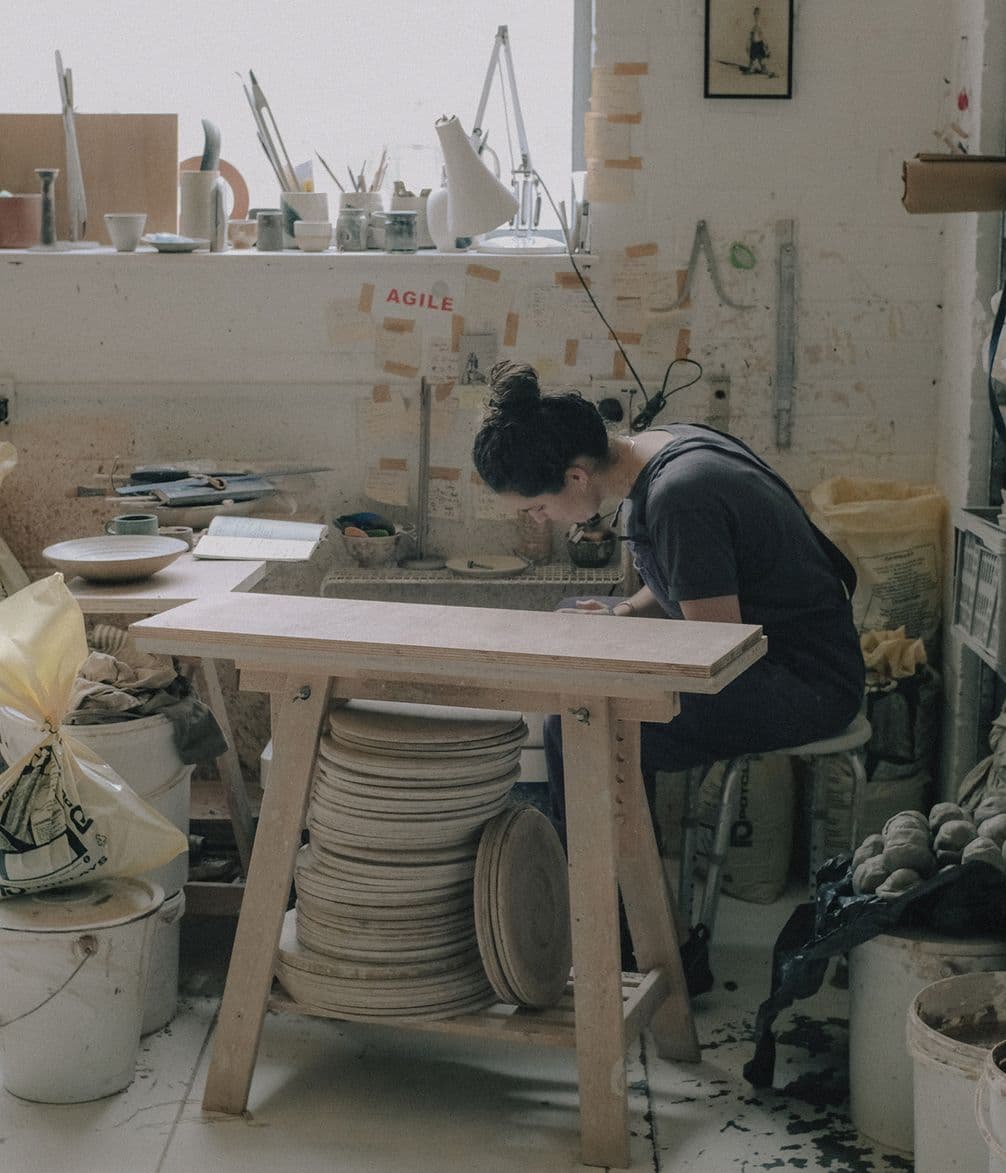 A person working intently at a pottery studio, surrounded by clay products, tools, and a cluttered workbench under natural light.