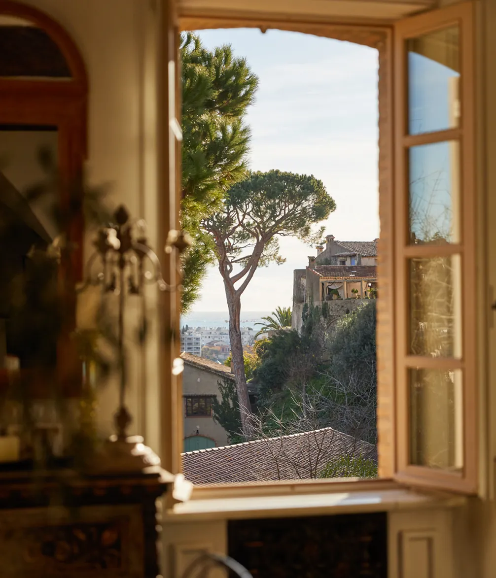 Open window view of a picturesque landscape with trees, rooftops, and distant ocean under a clear blue sky.