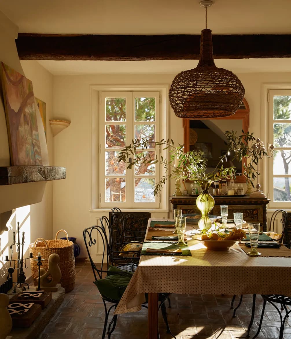 Sunlit dining room with a woven pendant light, rustic table set with glassware, and leafy plants. Sunlight filters through large windows.
