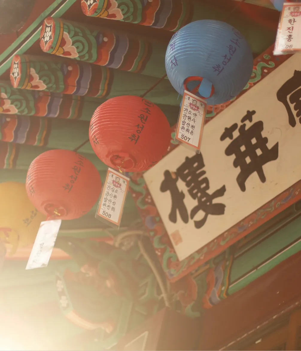 Colorful paper lanterns hang under an ornately painted wooden roof with Korean text, lit warmly by the sun.