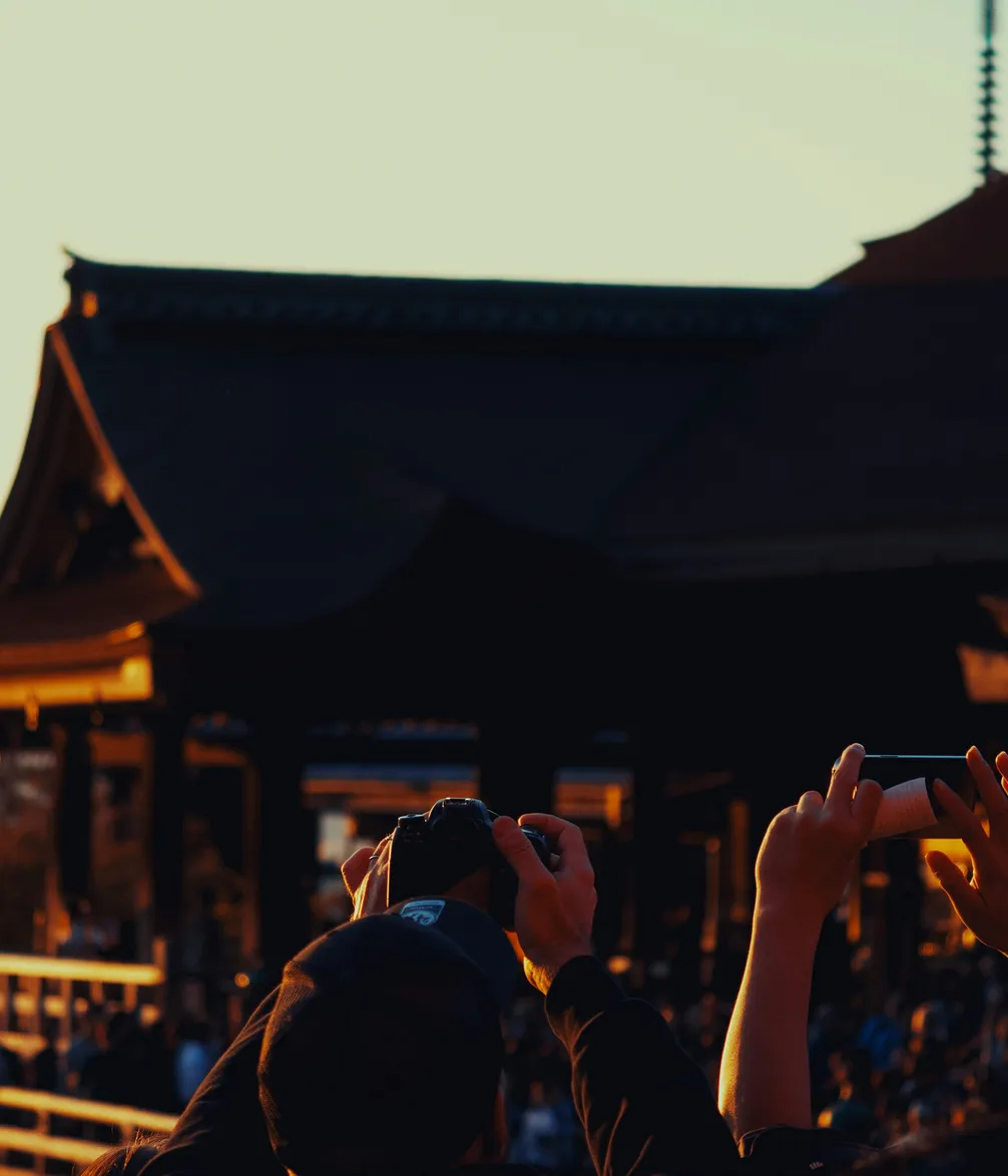 People taking photos with cameras and smartphones at sunset, with silhouettes of traditional architecture in the background.