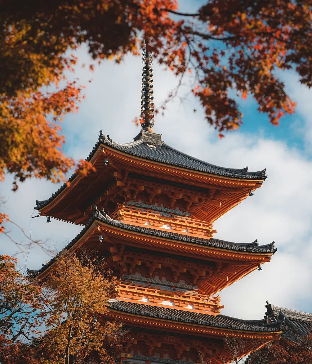 A traditional Japanese pagoda with red leaves in the foreground, set against a partly cloudy blue sky.