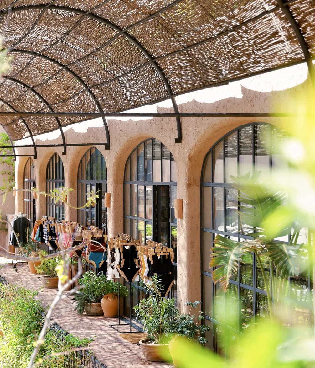 Outdoor clothing store display with hanging garments beneath an arched pergola, flanked by potted plants and large glass windows.