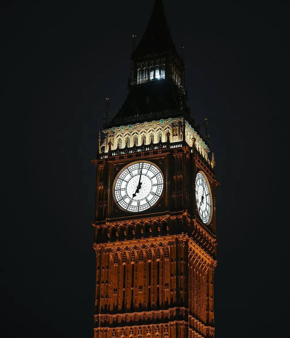 The illuminated clock tower of Big Ben at night, with glowing clock faces and ornate architectural details against a dark sky.