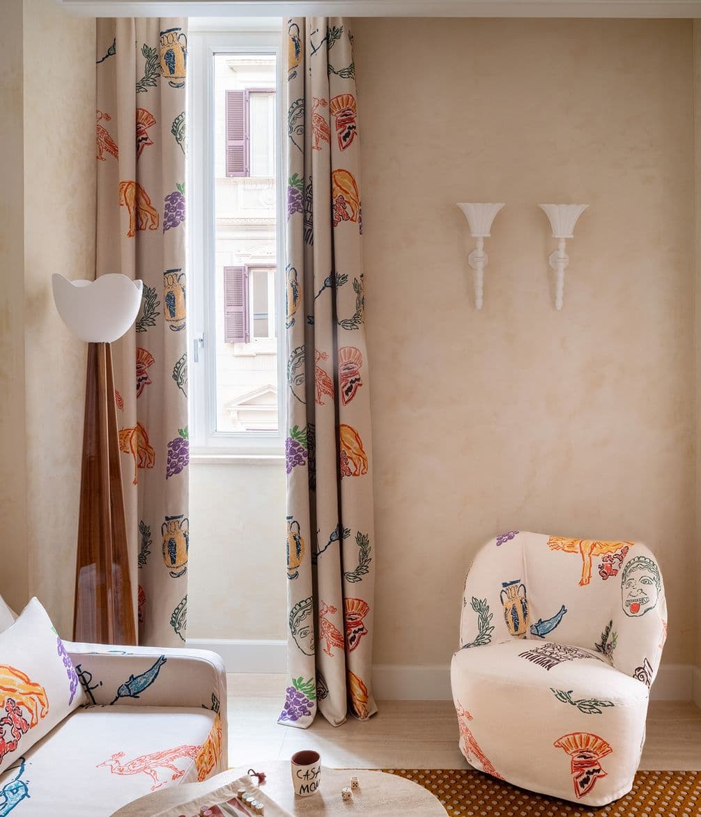 Room with colorful patterned curtains, matching chair, and sofa. Beige walls with two white sconces and light wooden flooring. Classic window view.