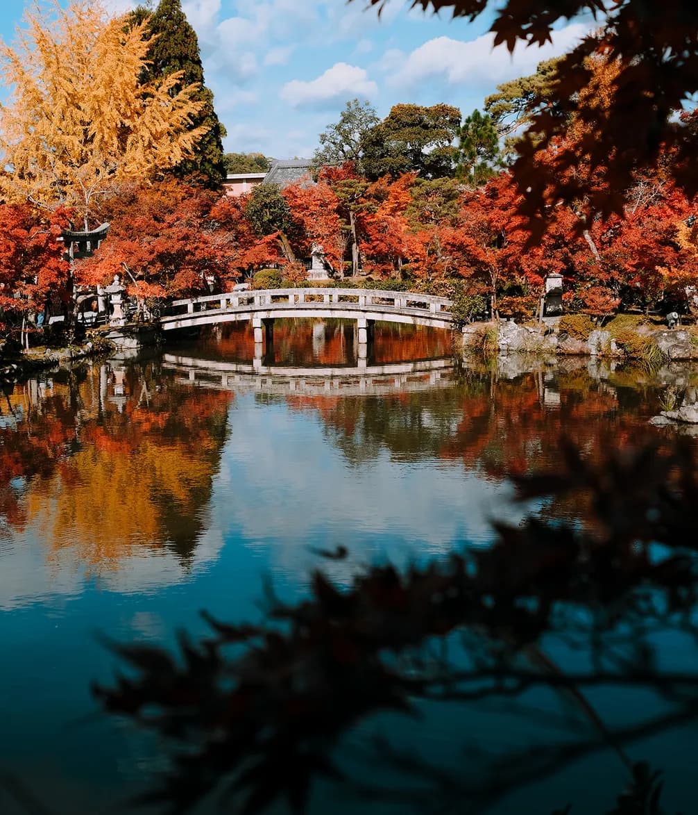 A serene pond reflects a stone bridge and vibrant autumn trees with red and yellow leaves under a blue sky.