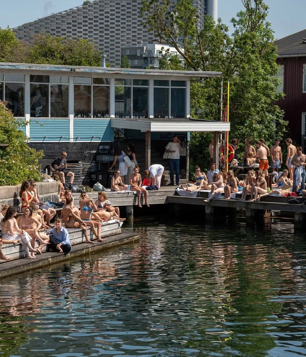 People sitting and sunbathing by the waterside on a sunny day, with a backdrop of trees and a building.