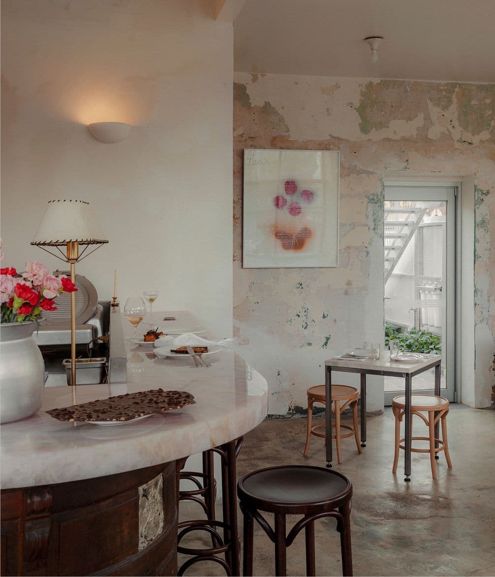 Cozy café interior with rustic walls, a small dining area, marble counter, stools, flowers, and soft lighting; artwork hangs on the wall.