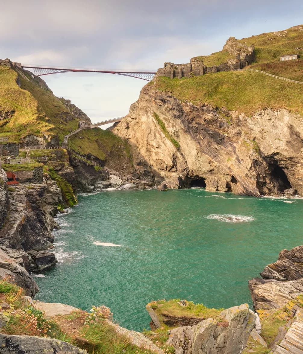 Coastal landscape with a turquoise bay, rocky cliffs, and a modern bridge connecting the cliffs. Rocky outcrops are covered with grass and plants.