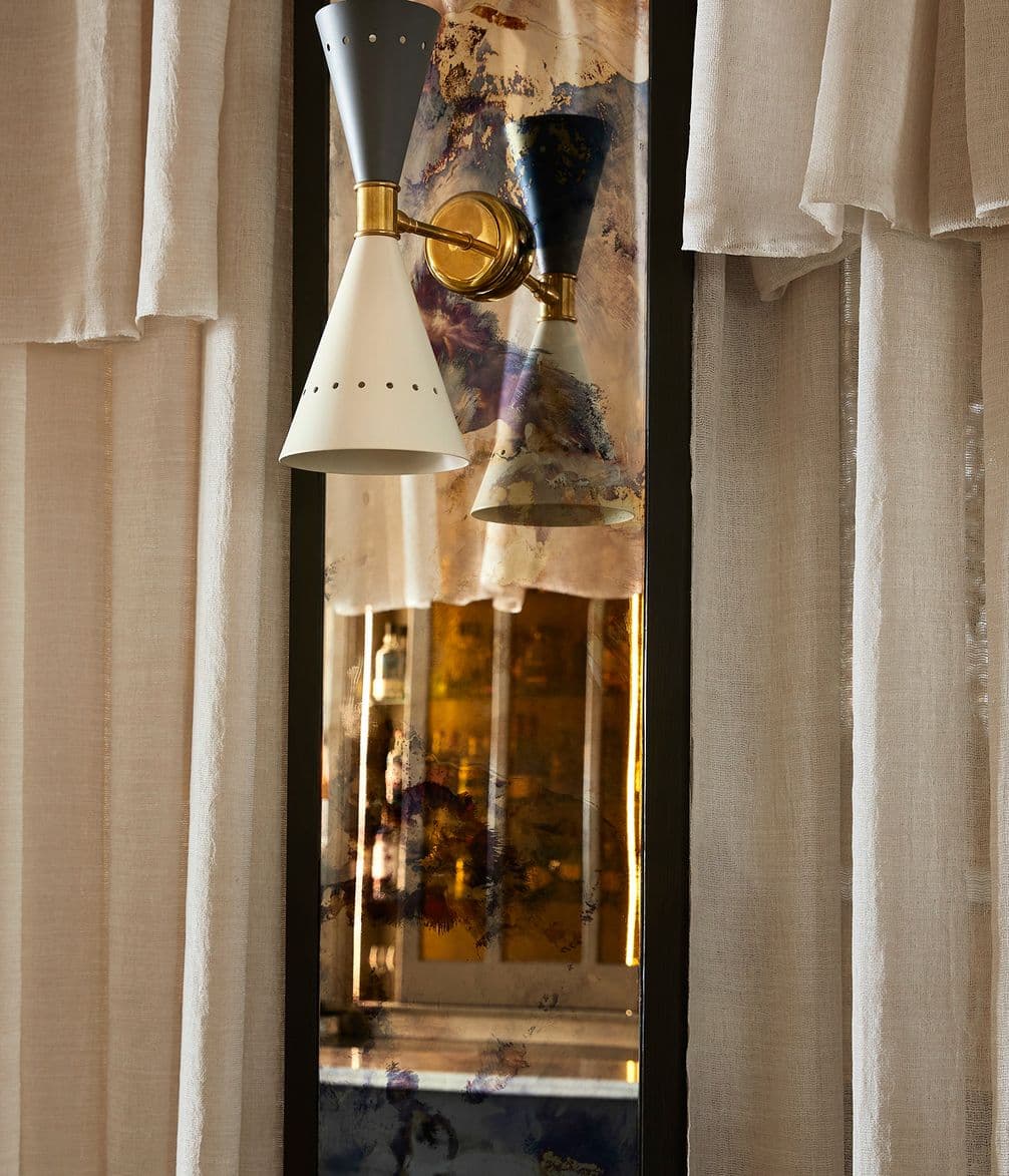 A modern wall sconce with a cone-shaped design is reflected in a tall mirror, surrounded by soft, cream-colored curtains.