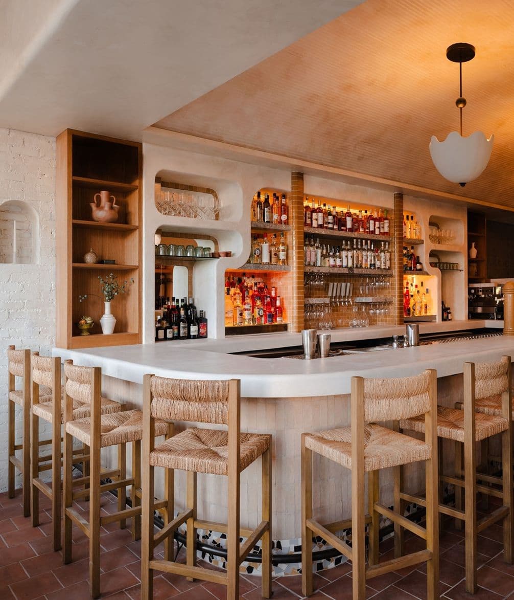 Stylish bar interior with high wooden stools, a curved white counter, and a variety of bottles on shelves, under warm ambient lighting.