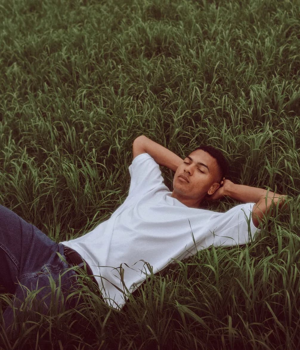 Person lying on green grass, wearing a white t-shirt and jeans, hands behind head, looking relaxed and peaceful.