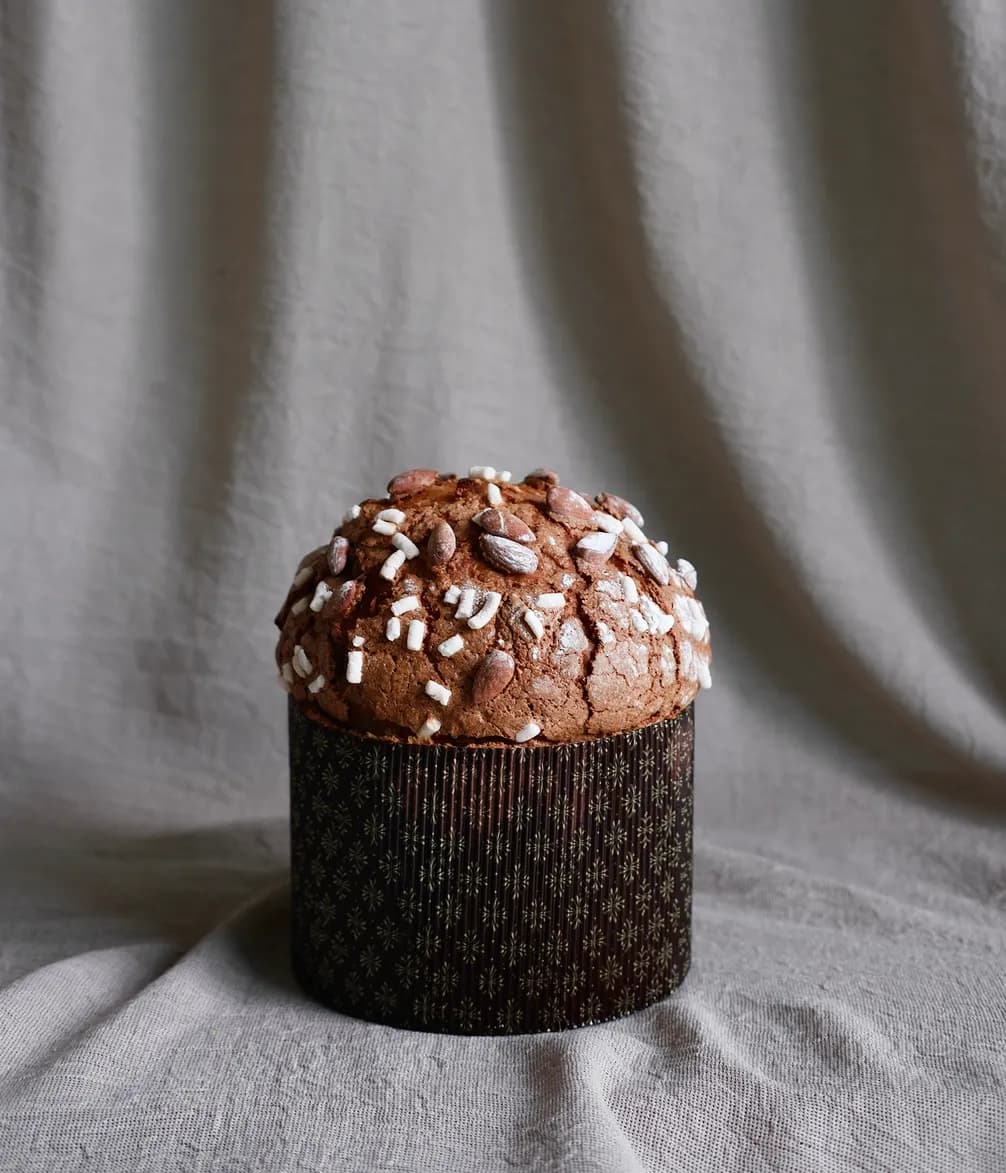 A tall, round Italian panettone with a golden-brown crust topped with almonds and sugar, set against a textured, muted background.