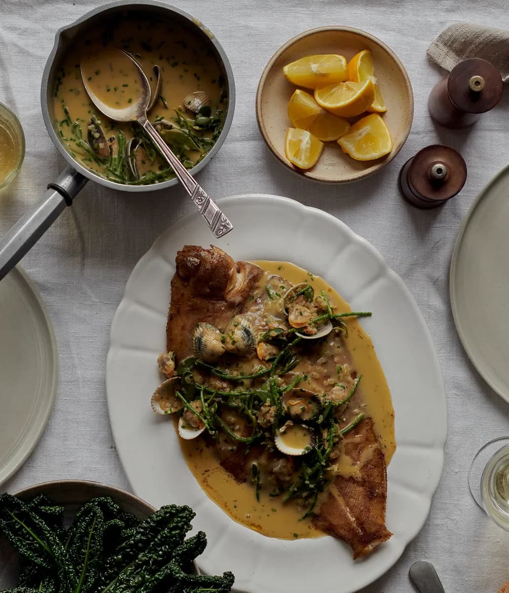 A plated whole fish topped with clams and sauce, accompanied by lemon wedges, sauce in a pan, salt and pepper shakers, and greens on a cloth-covered table.