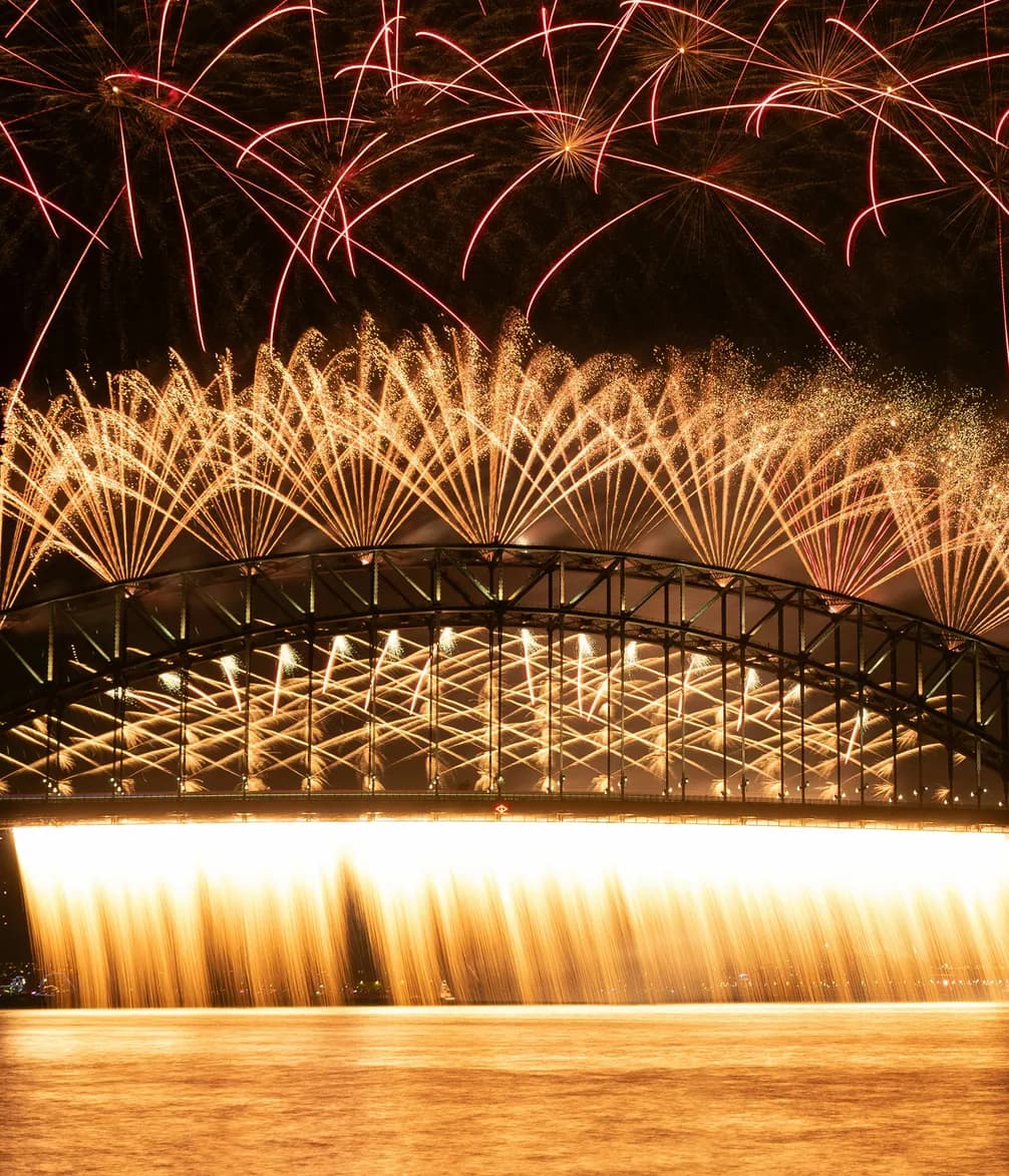 Fireworks display over a large, illuminated bridge with vibrant red and gold bursts lighting up the night sky, reflecting on the water.