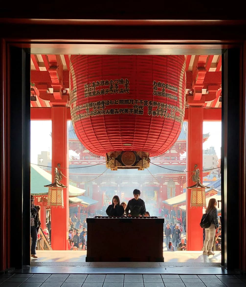 People stand before a large red lantern at the entrance of a bustling temple with vibrant colors and intricate architecture.