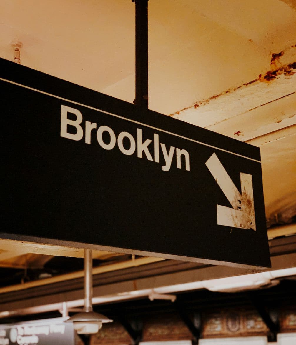 A subway sign with "Brooklyn" and a downward arrow, located in a dimly lit underground station with a rustic ceiling.
