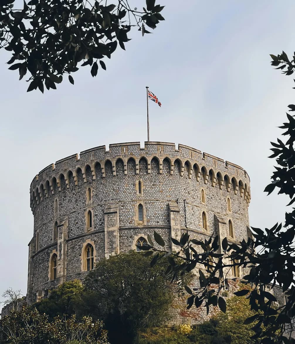 A round stone castle tower with a Union Jack flag atop, framed by branches and leaves, under a clear sky.