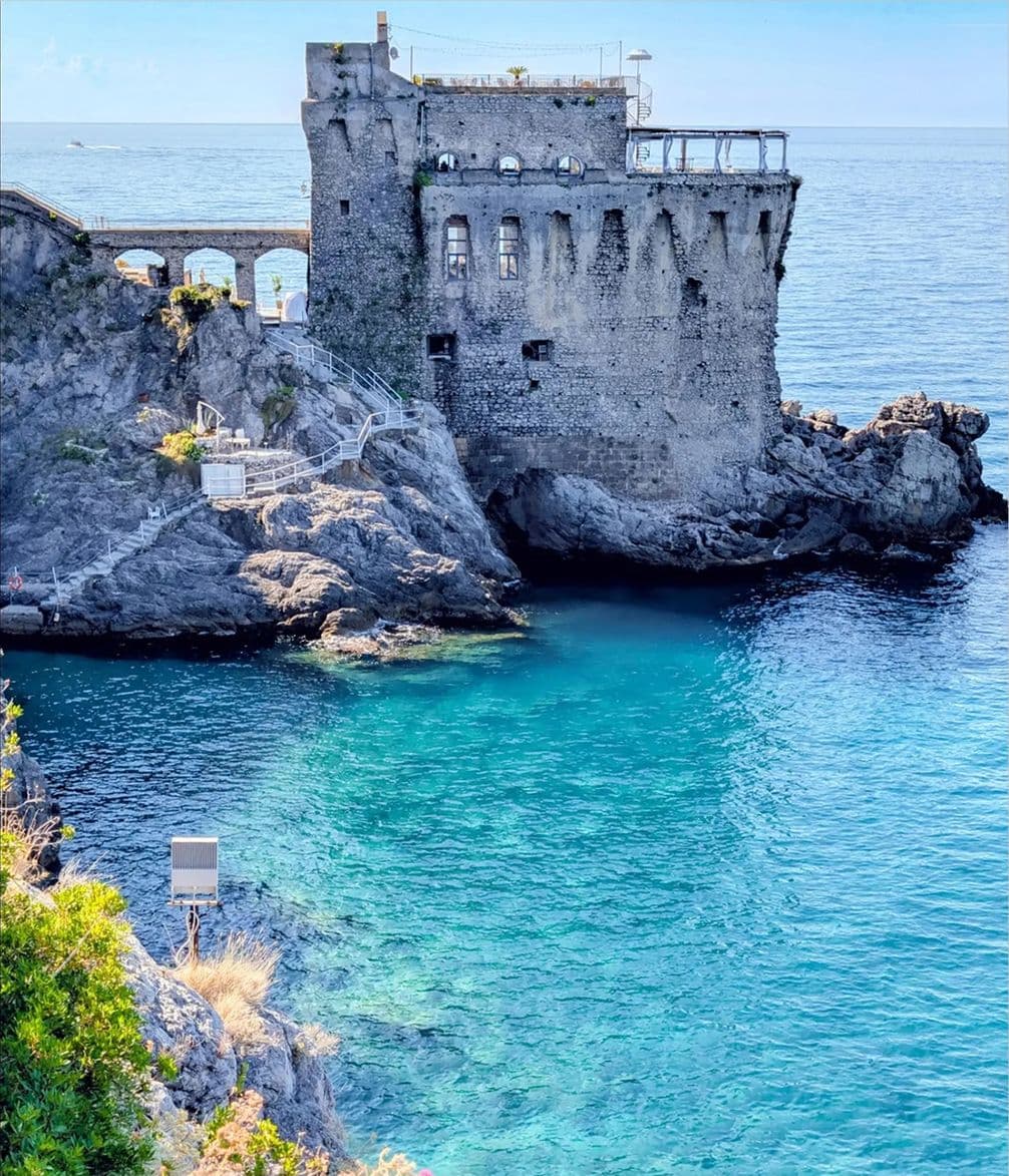 Historic stone tower perched on a rocky coast, surrounded by clear turquoise water under a bright blue sky.