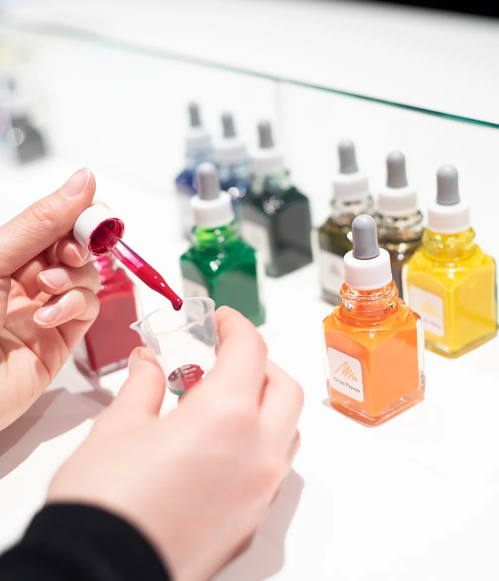 Hands use a dropper to add red liquid to a small measuring cup, with many colorful dropper bottles in the blurry background.