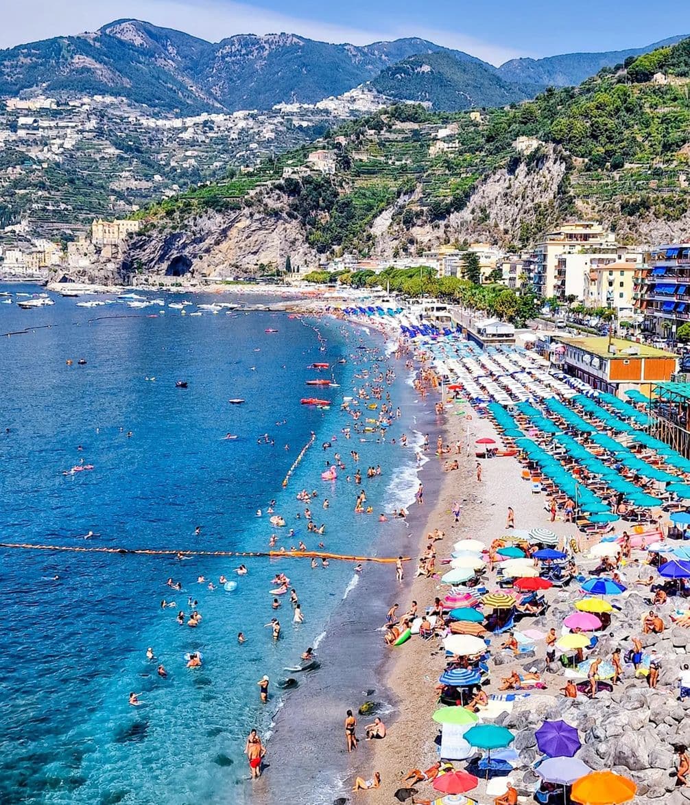 A vibrant beachfront with colorful umbrellas and sunbathers, blue waters, and scenic mountains in the background.