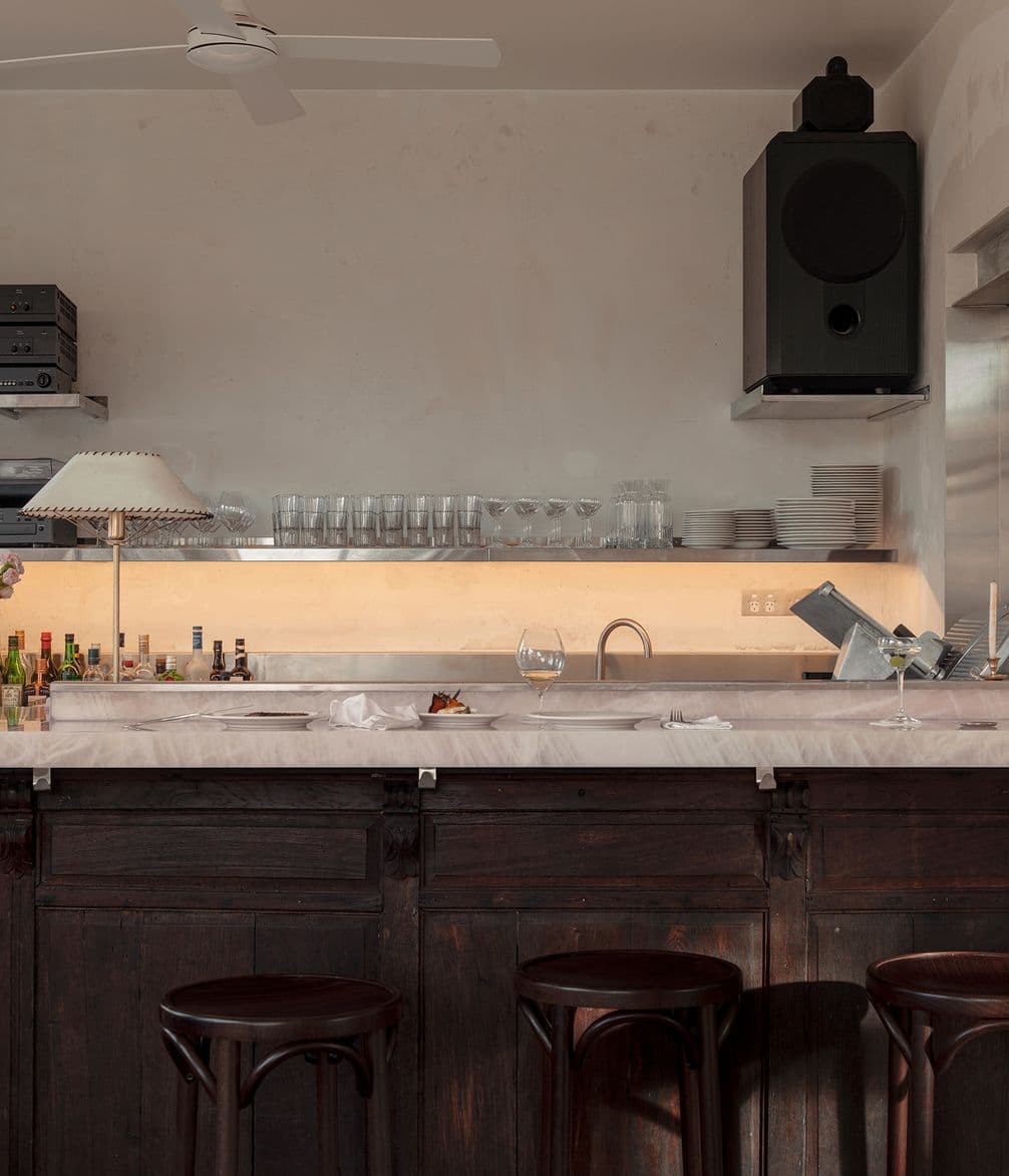 Modern kitchen with dark wood cabinets, marble countertop, bar stools, hanging speaker, glassware on shelves, and a countertop lamp.