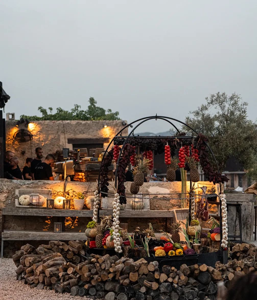 Outdoor rustic market stall with hanging garlic, tomatoes, and chili peppers, surrounded by stacks of firewood and lit by warm lanterns.