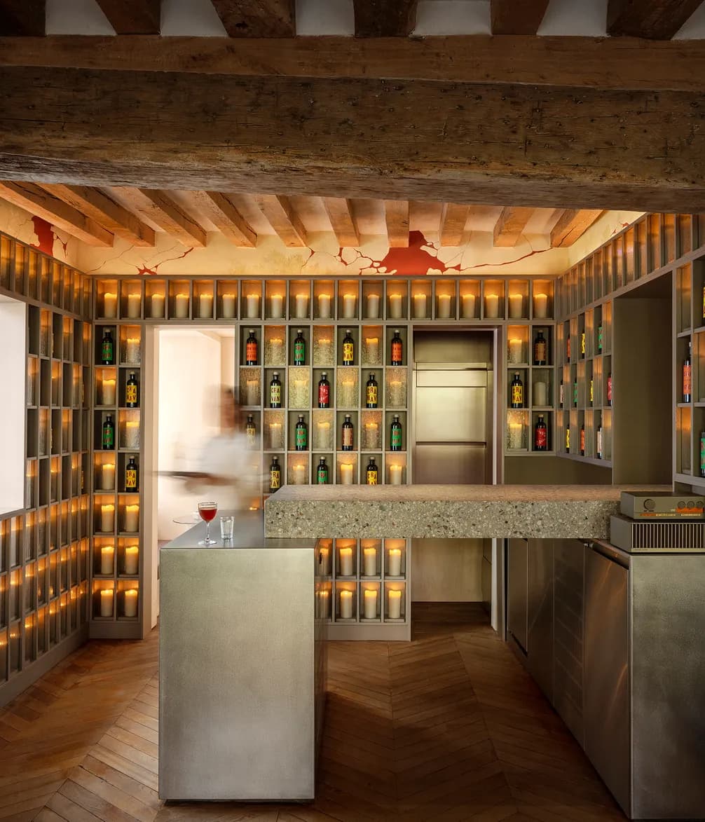 A modern bar with wooden shelves filled with colorful bottles, lit candles below, and a granite counter on a herringbone wood floor.
