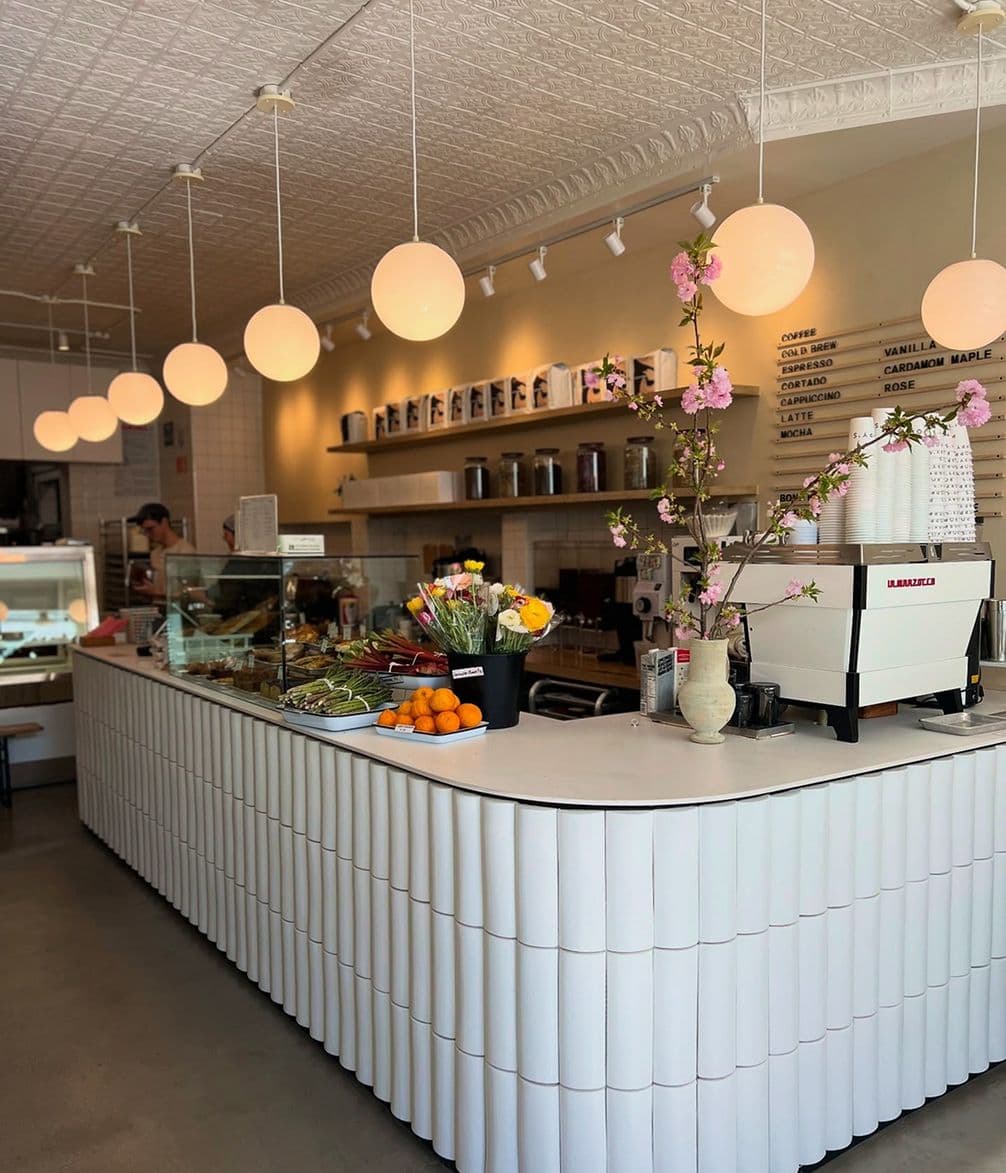 Contemporary café with a white counter, hanging globe lights, flowers, oranges, and a coffee machine; menu board on the wall.