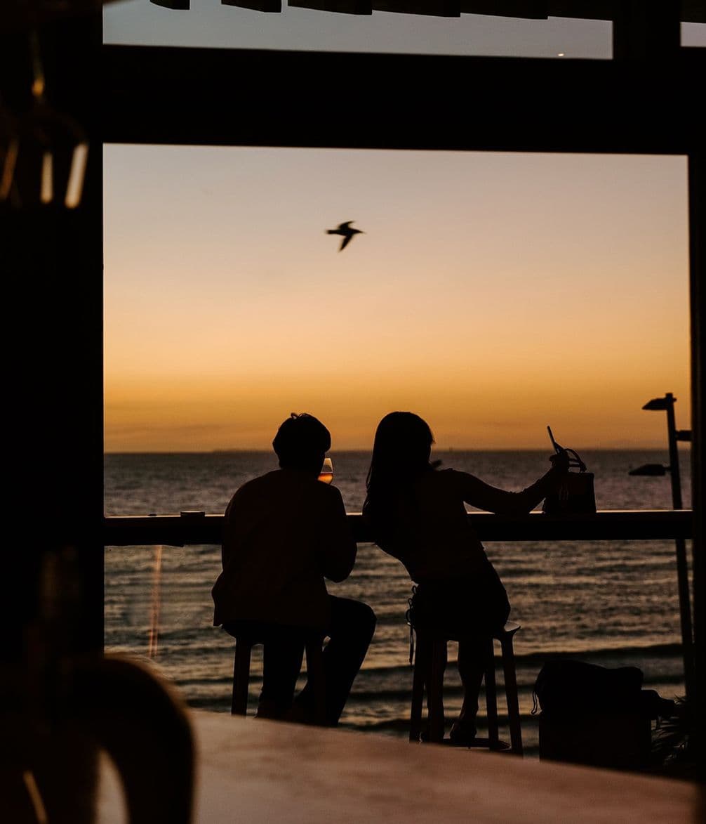 A couple sits at a bar, silhouetted against a vibrant sunset over the ocean, with a bird flying in the sky.