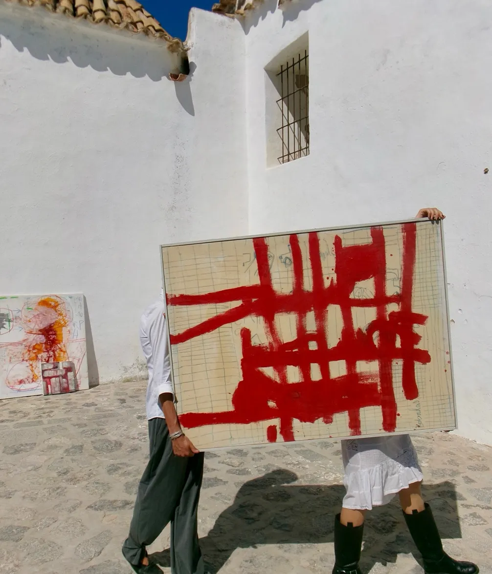 Two people carrying a large abstract painting with red brushstrokes, in front of a white building under a clear blue sky.