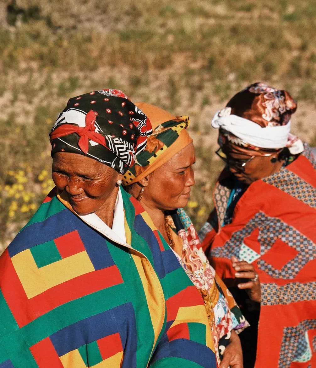 Three women wearing colorful, patterned wraps and headscarves stand outdoors, with vibrant geometric designs and lush greenery in the background.